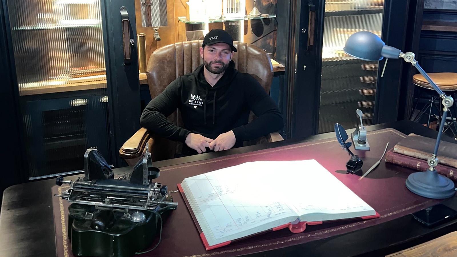 Rob Prager sits in a leather chair behind a large desk. He is smiling and wearing a black baseball cap and a black hoodie.