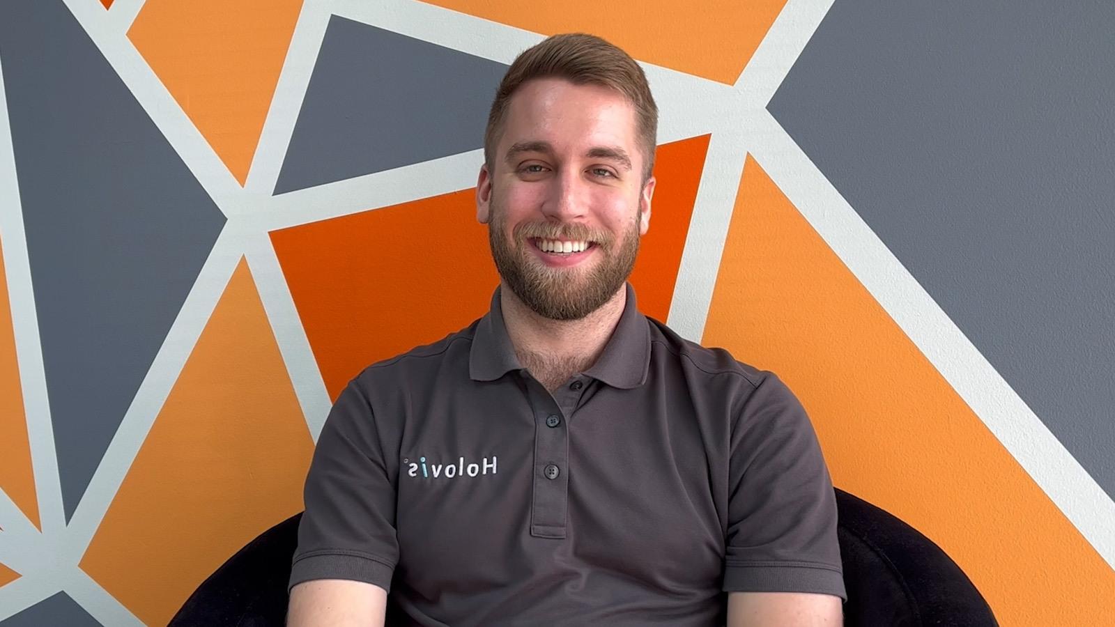 A man with light hair and a beard wearing a grey Holovis polo while seated and smiling in front of an orange, grey, and white geometric background.