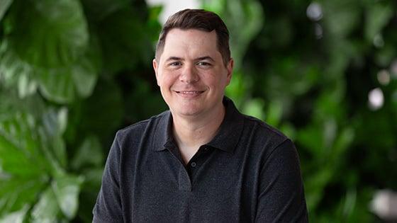 A man with short brown hair is wearing a dark grey polo while smiling against a lush green backdrop.
