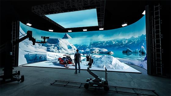 一个穿着牛仔裤、运动鞋和格子衬衫的男人站在制作工作室里. 他面对着一个巨大的LED屏幕，屏幕上显示着冰封的山脉和雪地摩托.