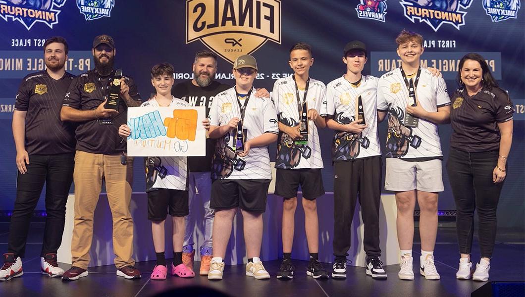 A group of players and coaches in esports jerseys on stage, one player holds a sign that reads “For Hank.”